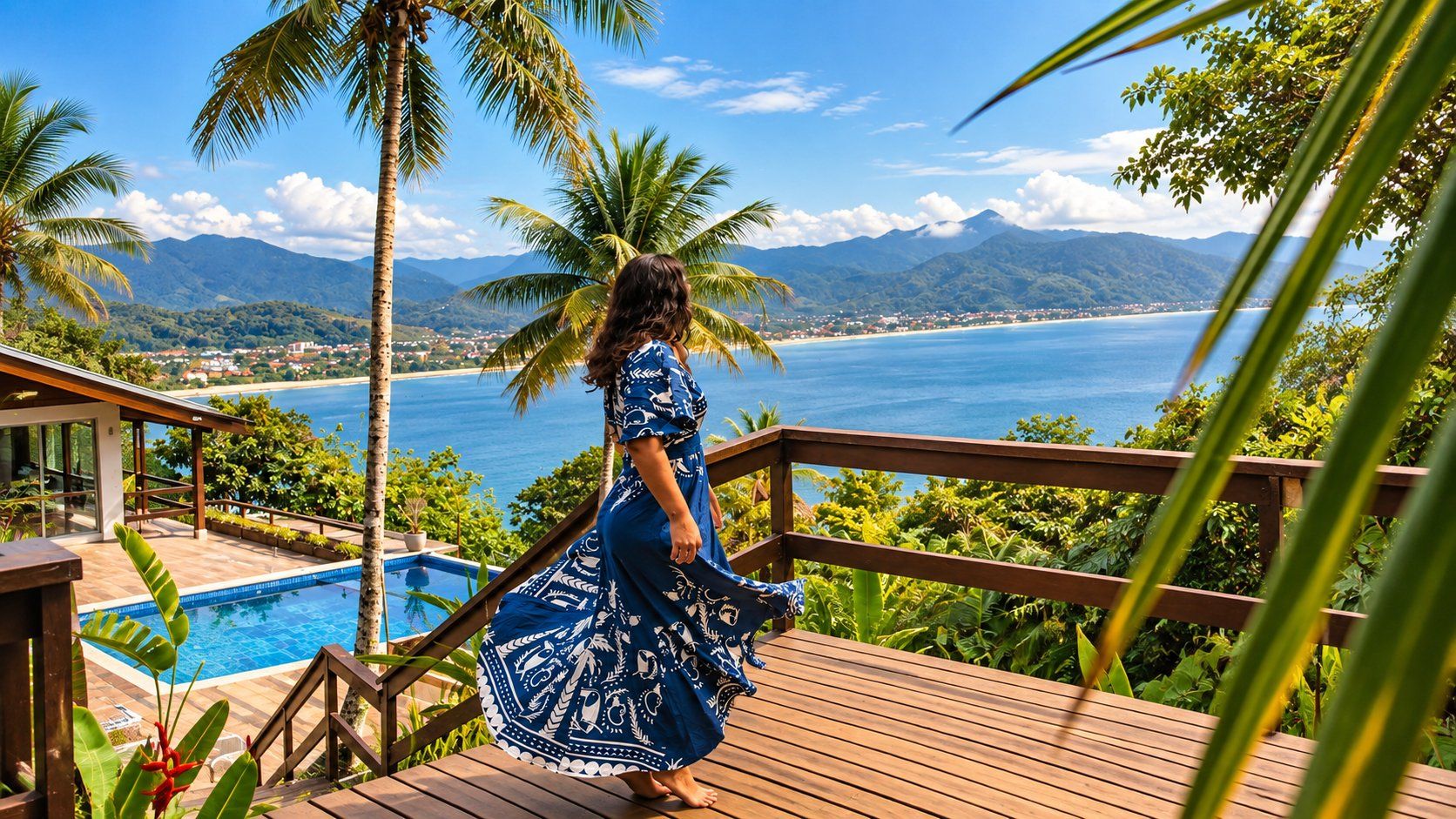 A woman in a blue dress standing on a wooden deck overlooking a pool and a coastal view with mountains in the distance.