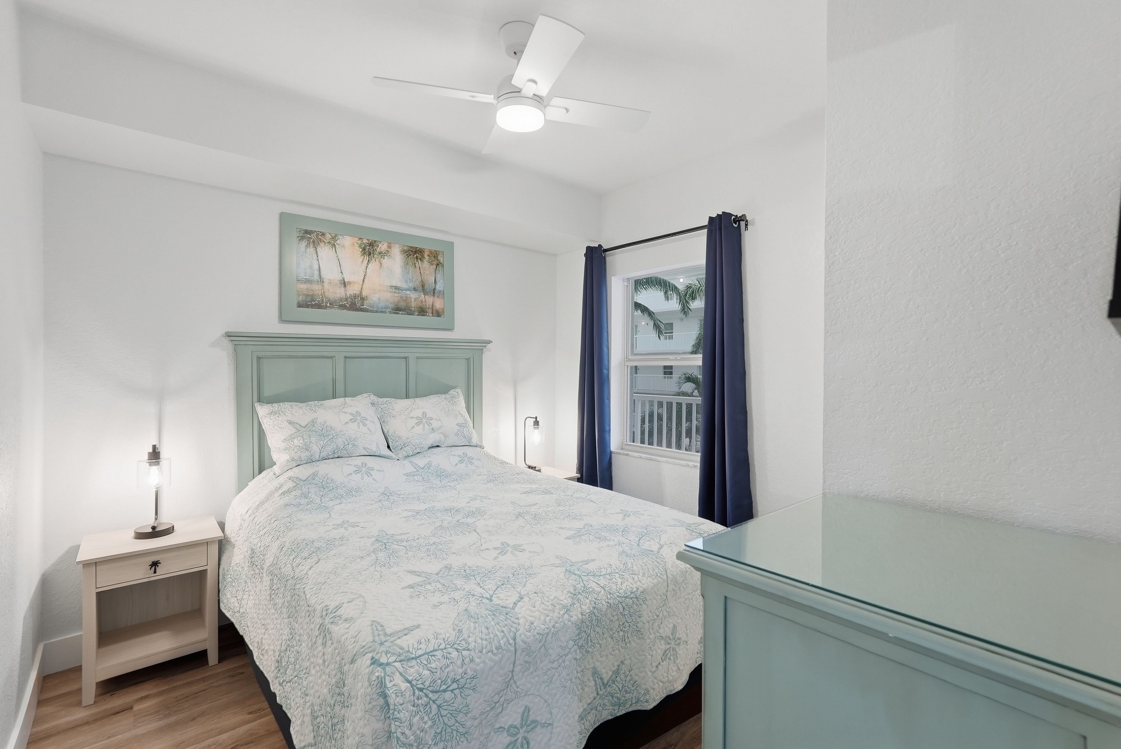 A bedroom with a bed, a nightstand, a desk, a ceiling fan, a window with curtains, and a painting on the wall.