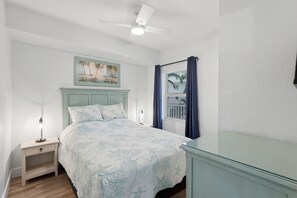 A bedroom with a bed, a nightstand, a desk, a ceiling fan, a window with curtains, and a painting on the wall. - Elevated Beach Escape on Treasure Island (Treasure Island)