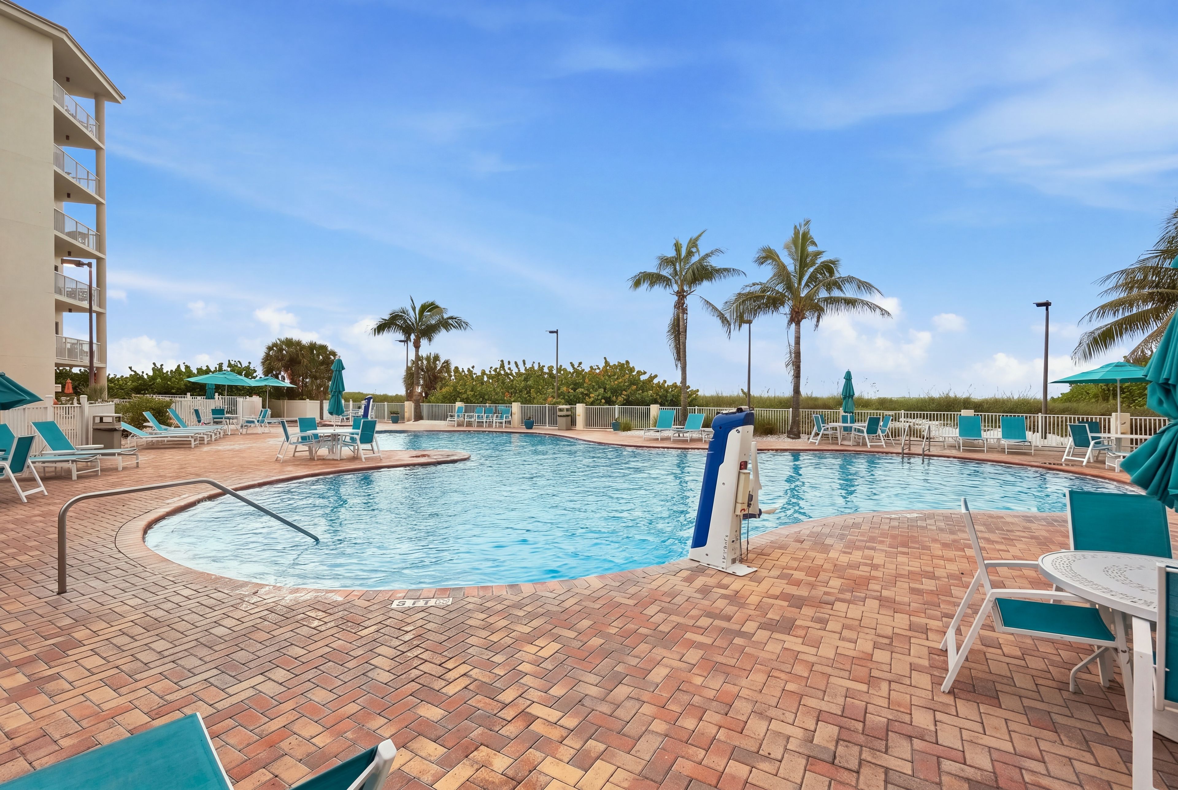 A pool area with lounge chairs and umbrellas, surrounded by palm trees and a clear sky.