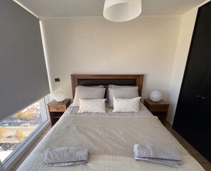 A bedroom with a large bed, two bedside tables, a headboard, and a view through a window. - Edificio Level by Domus Travels (Ñuñoa)