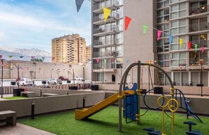 Ein Spielplatz im Freien mit Rutsche und Klettergerüst, umgeben von einem Gebäude mit Balkonen und einem Parkplatz.