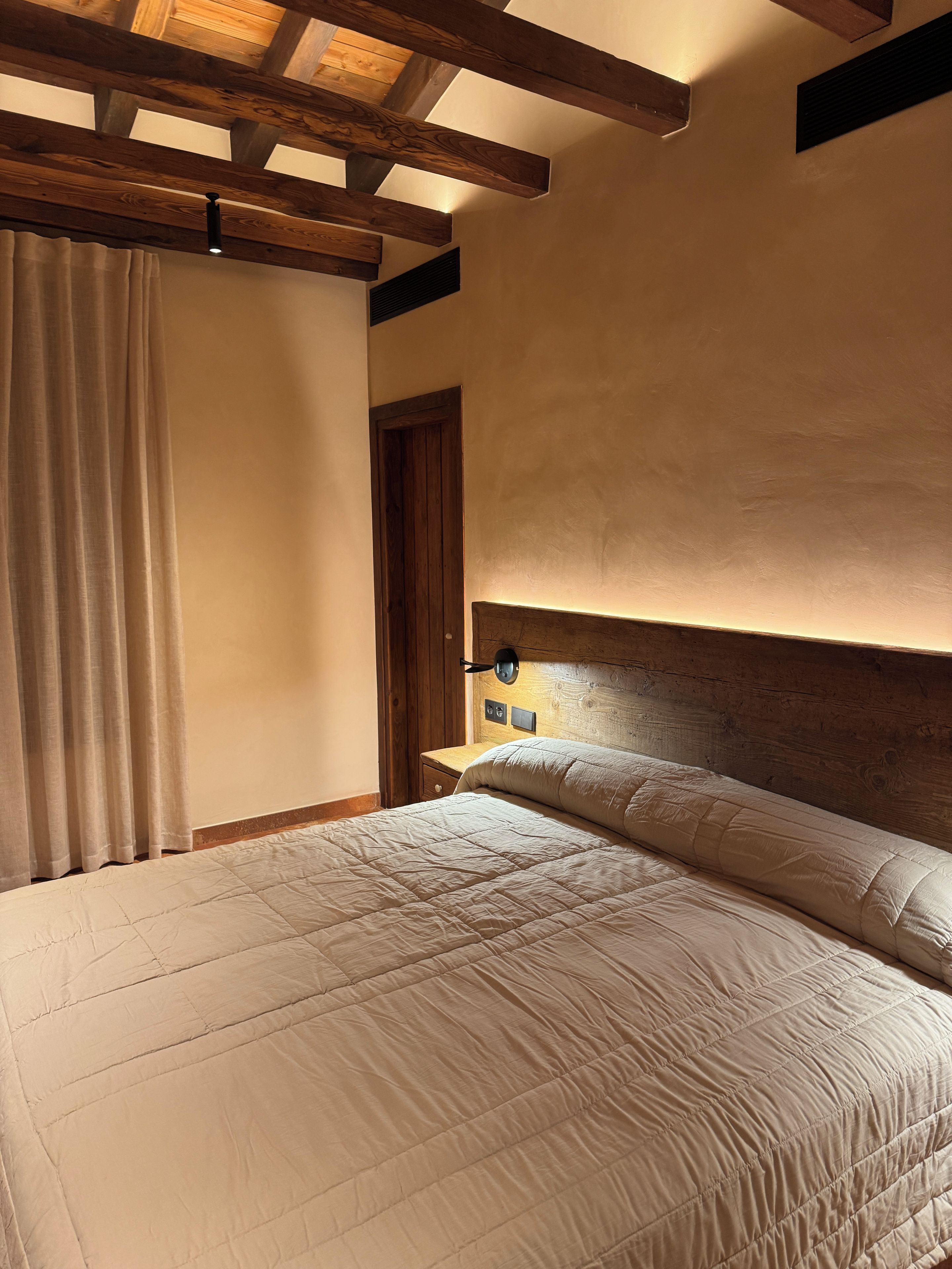 Un dormitorio con techo de madera, una cama con cabecera, una mesita de noche y una lámpara de pared.