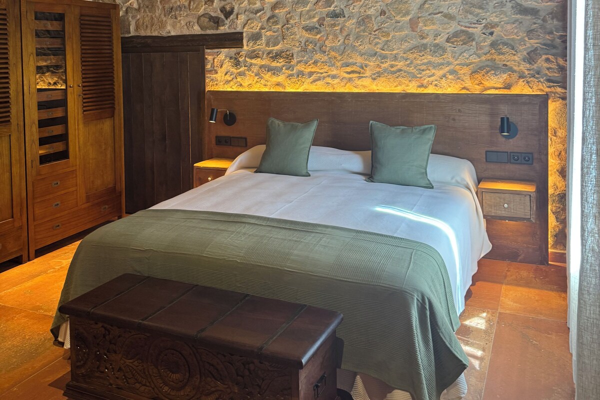 Una camera da letto con parete in pietra, testiera in legno, un letto con biancheria verde, un comodino e un settimino in legno.