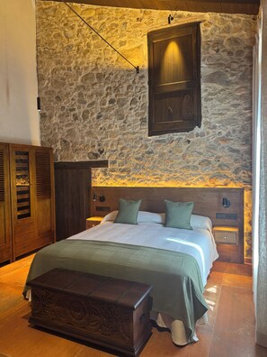 Una camera da letto con parete in pietra, testiera in legno, un letto con biancheria verde, un comodino e un settimino in legno.