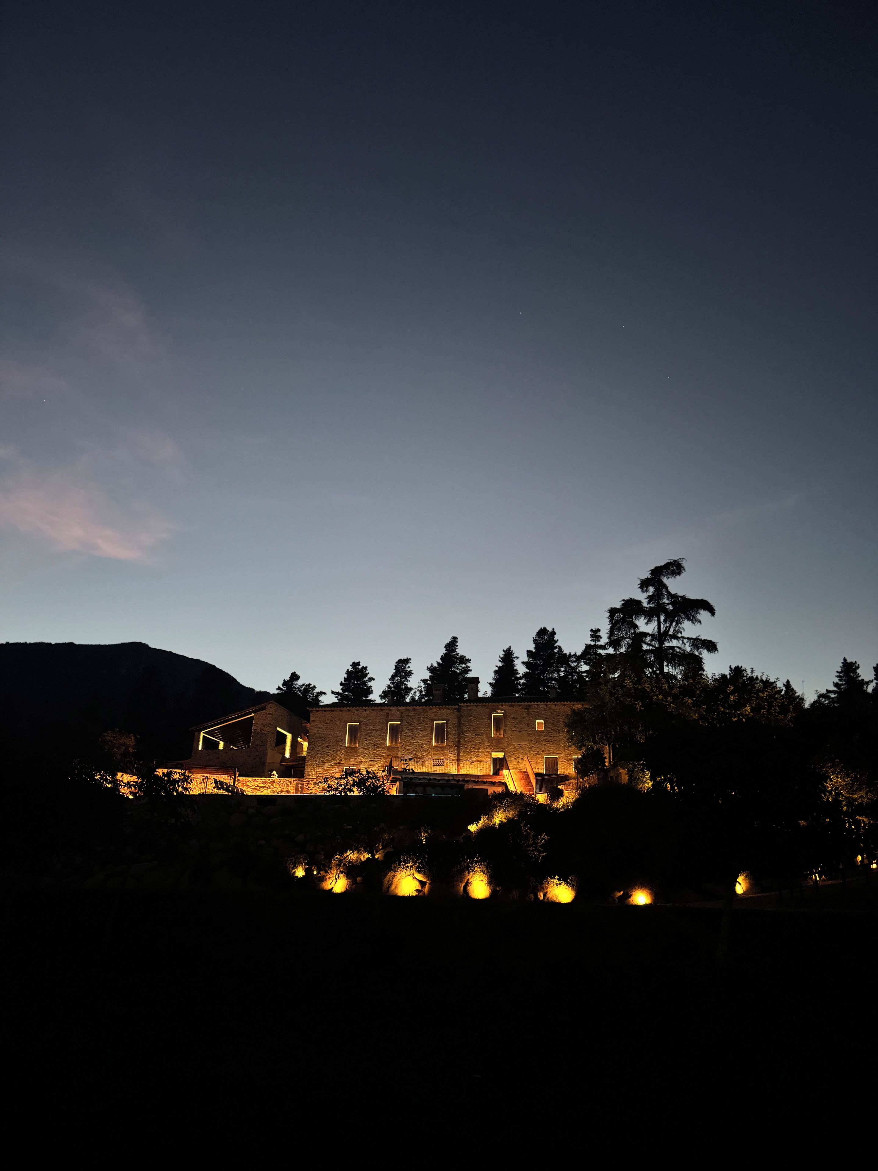 A building with multiple windows and a stone facade, surrounded by trees and hills.