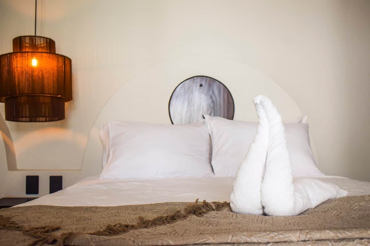 Un lit avec des oreillers blancs et une couverture, un miroir rond accroché au mur et un luminaire suspendu.