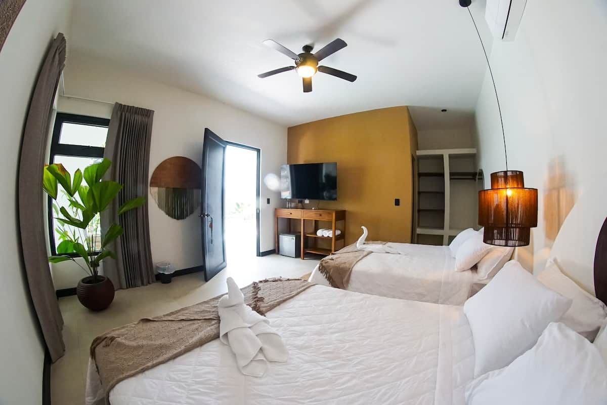 Quarto de hotel com duas camas, ventilador de teto, televisão, planta e abajur.