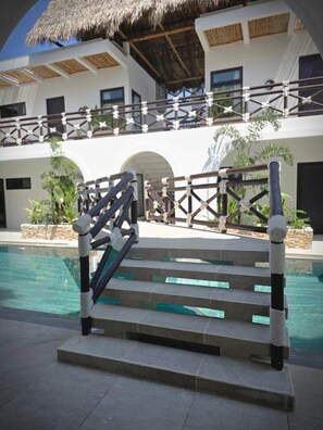 Área de piscina com escadaria para um balcão, cercada por um edifício branco com telhado de palha.
