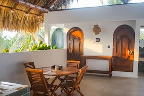 A dining area with a round table and chairs, a thatched roof, and a view of palm trees.