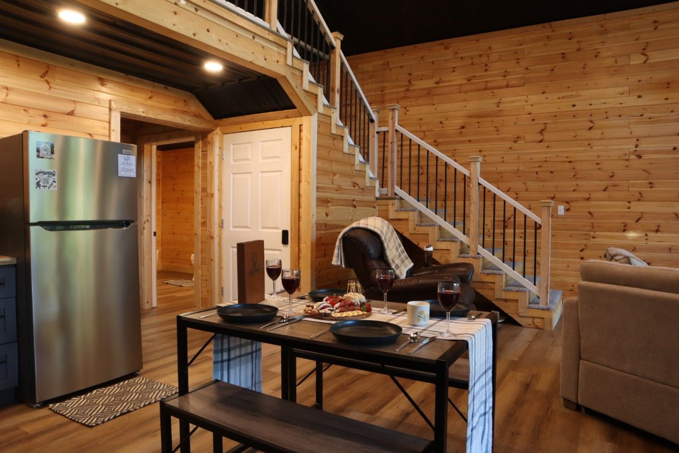 A wooden cabin interior with a dining table set for two, a refrigerator, and a staircase.