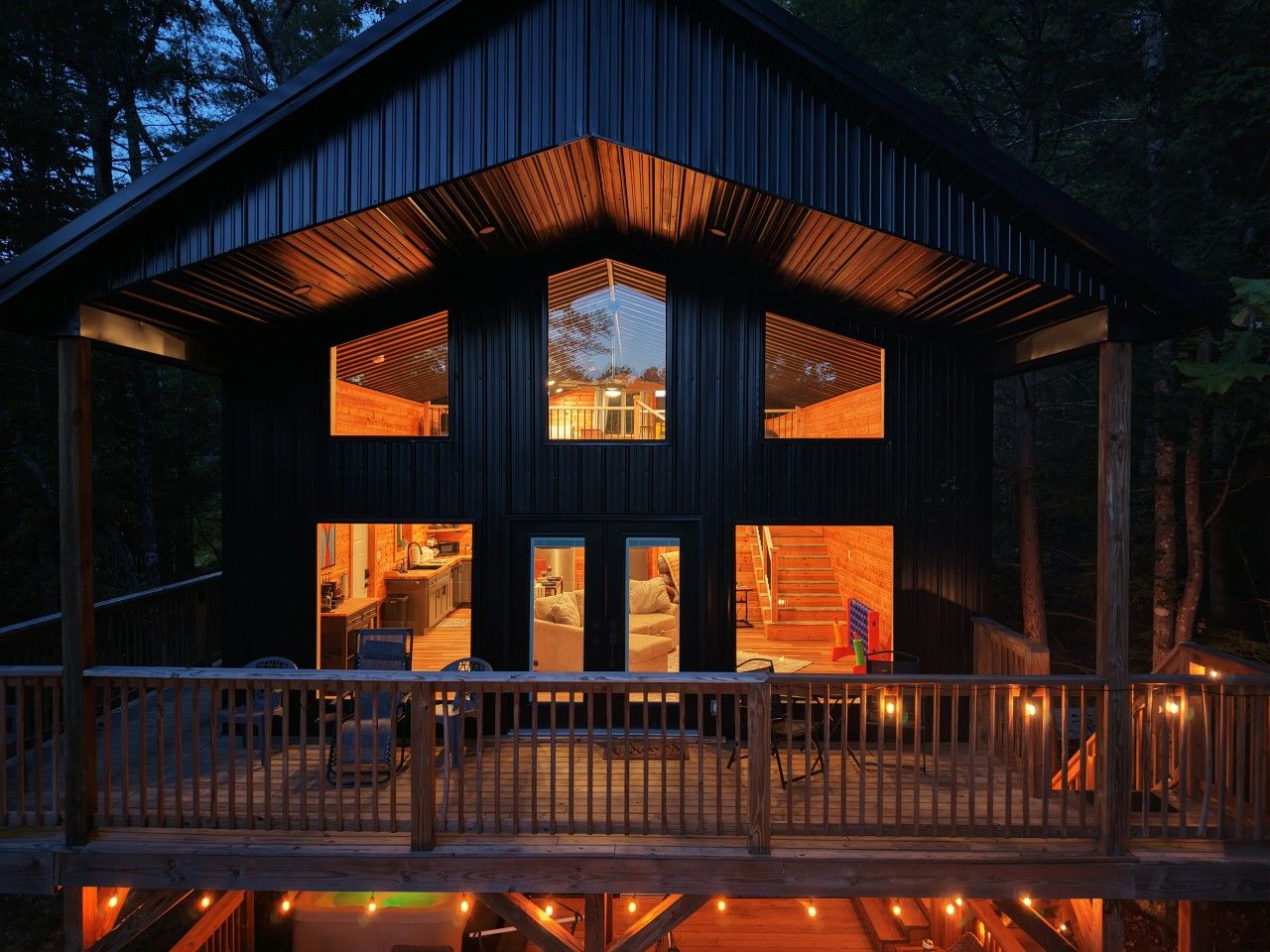 A two-story cabin with a deck, wooden railings, and interior lighting.