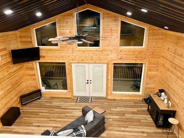 A wooden cabin with a loft bedroom, a sofa, a TV, and a ceiling fan. - Chasin Turkeys | Lakefront Cabin w/ Hot Tub, Arcade Game & Fire Pit (Campton)
