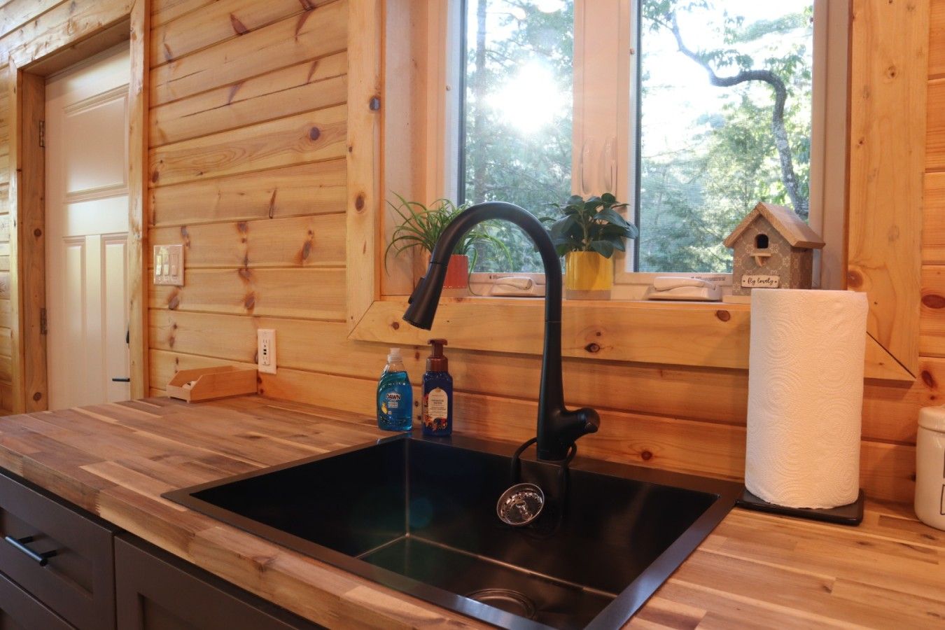 A wooden cabin interior with a black sink, wooden cabinets, a window with a view of trees, and a small birdhouse on the windowsill.