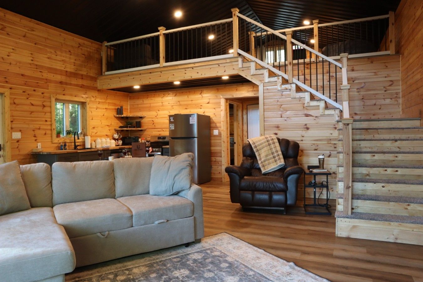 A log cabin interior with a sofa, armchair, and a staircase leading to an upper level.