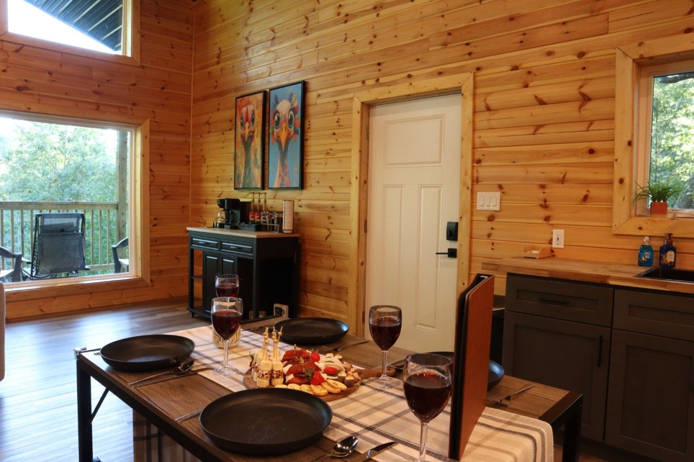 A wooden cabin interior with a dining table set for two, two framed paintings on the wall, a kitchen area with a sink, and a door leading to another room.