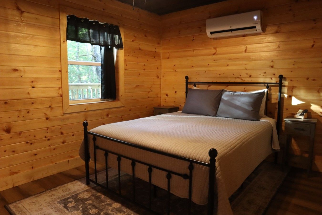 A wooden cabin bedroom with a bed, a window with curtains, an air conditioning unit, and a bedside table with a lamp.