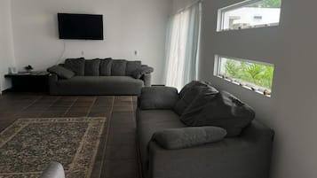 Sala de estar com sofá, TV na parede e janela com vista para o verde.