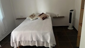 Una cama individual con una colcha blanca, dos almohadas, una mesita de noche y un calentador portátil.