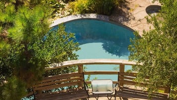 A swimming pool with a wooden deck and chairs, surrounded by greenery.