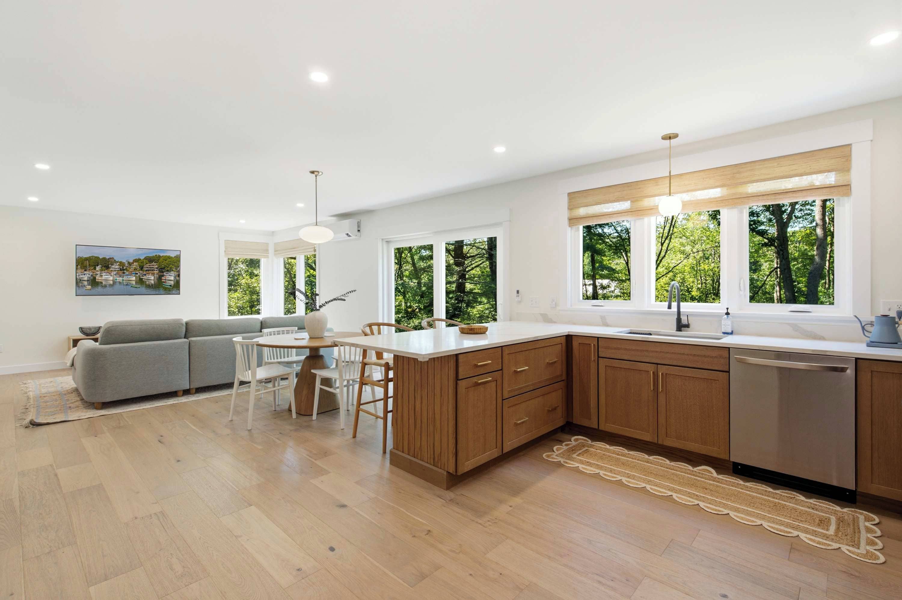 A modern kitchen with wooden cabinets, a dining area with a gray sofa, and large windows offering a view of trees.