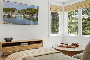Una habitación con un ventanal grande, un cuadro de una escena de lago, una consola de madera y una mesa redonda con una presentación de comida.