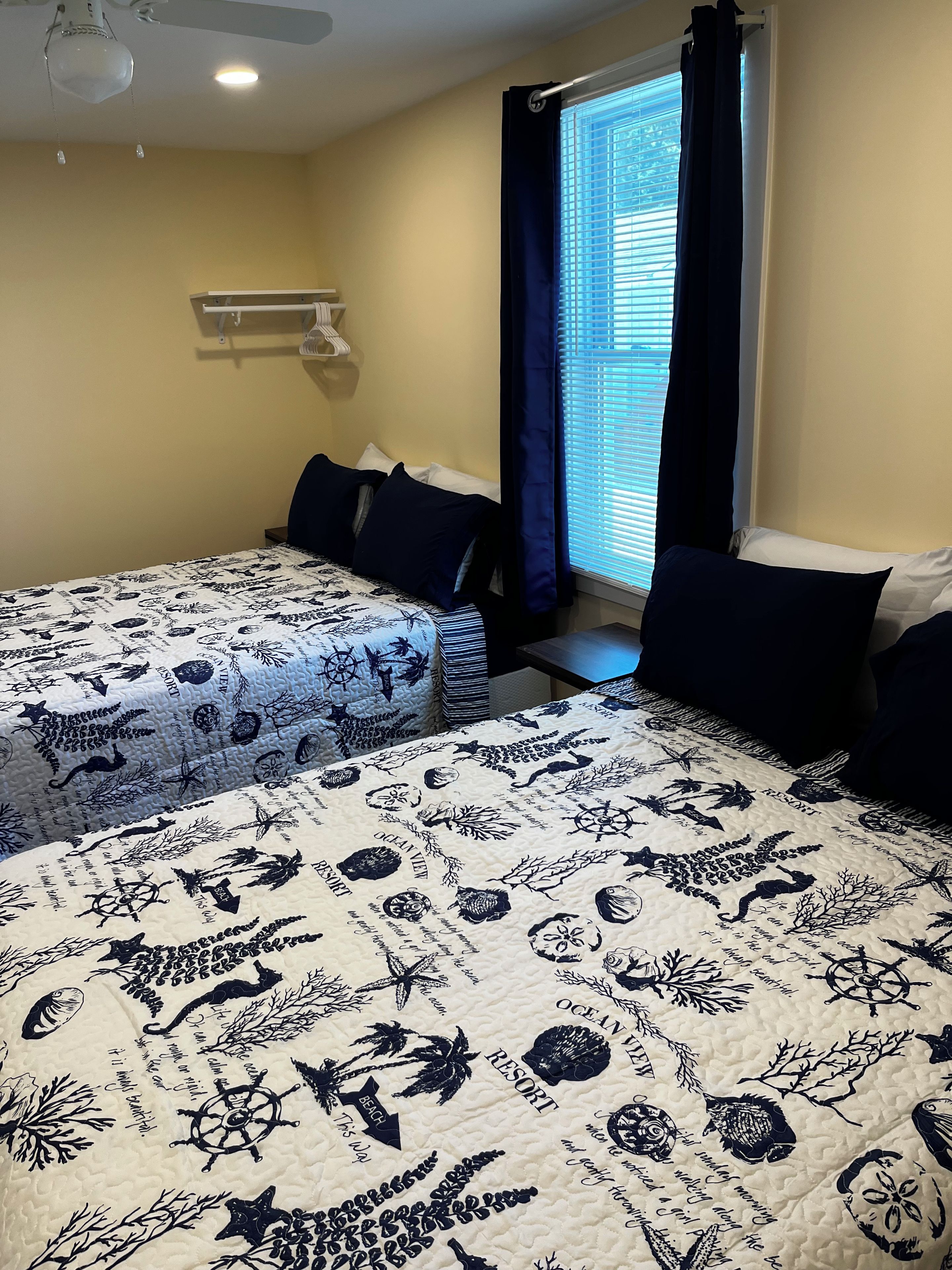 Ein Schlafzimmer mit zwei Betten, einem Fenster mit Jalousien, einem Deckenventilator und einem an der Wand befestigten Regal.