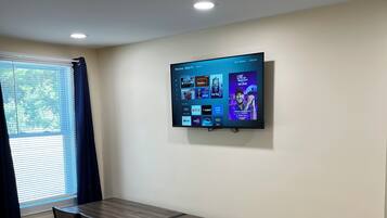 Ein Essbereich mit einem langen Tisch und Stühlen, einem flachbildfernseher an der Wand und einem Fenster mit Jalousien.
