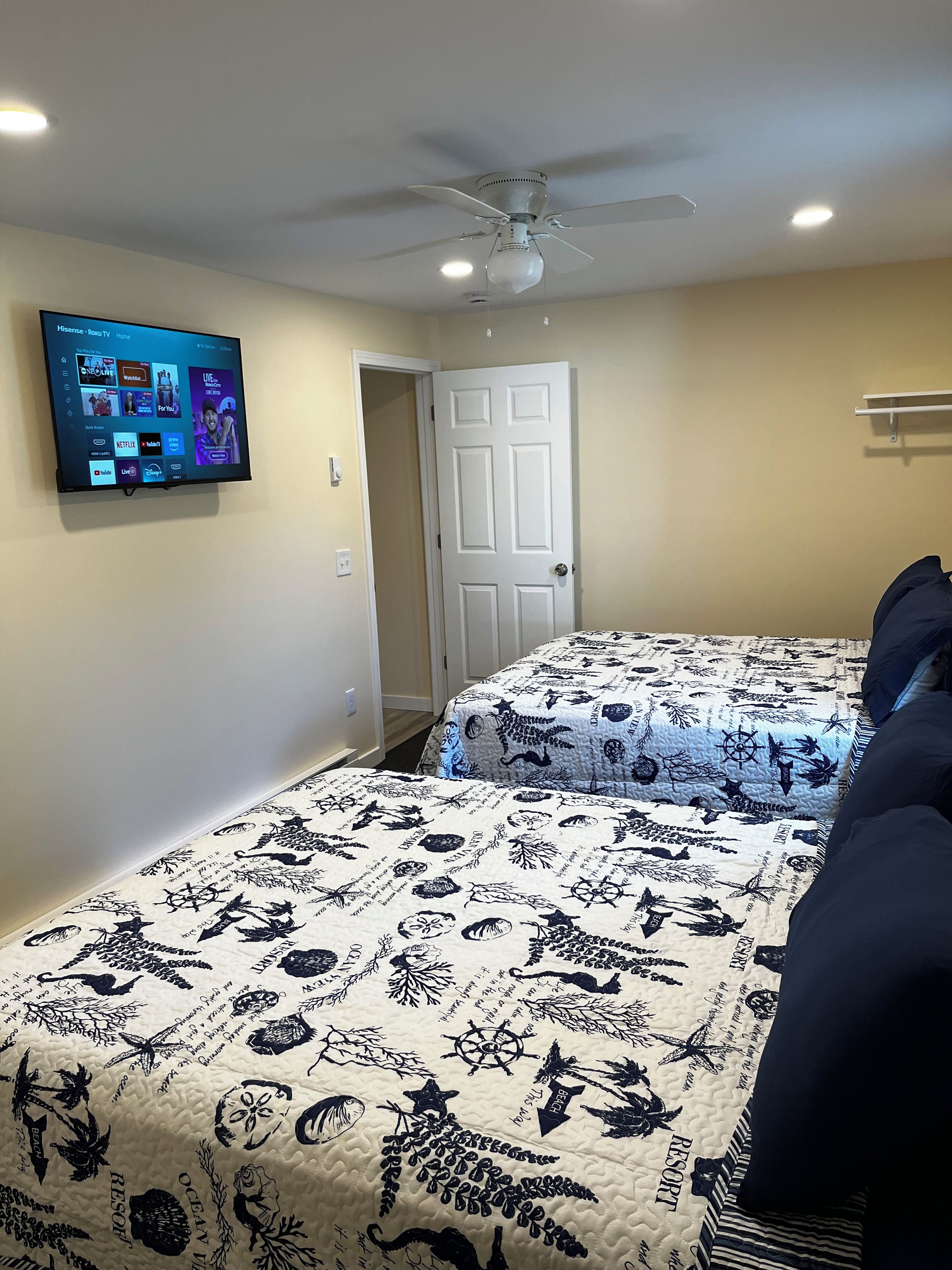 Un dormitorio con dos camas, ventilador de techo, televisor montado que muestra servicios de transmisión, y una puerta que da a otra habitación.