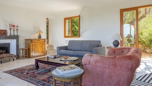 Ein Wohnzimmer mit Kamin, einer Couch, einem rosa Sessel und einem großen Fenster mit Blick auf Bäume.