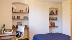 Ein Zimmer mit zwei hölzernen Regalen, gefüllt mit Dekoration, einem Stuhl mit gemustertem Sitzkissen und einem Bett mit blauer Bettdecke.