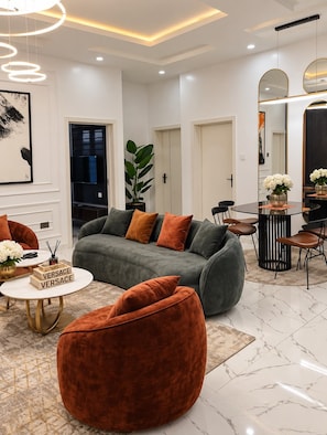 A modern living room with a grey sofa, orange pouf, and a round coffee table. There is a dining area with a black table and chairs, and a large mirror on the wall. - Nigus House Akinloye (Lekki)