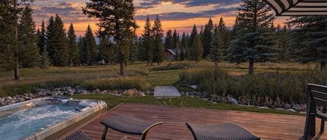 Un terrazzo in legno con vasca idromassaggio e poltrone relax, immerso in una foresta e con vista sul tramonto.