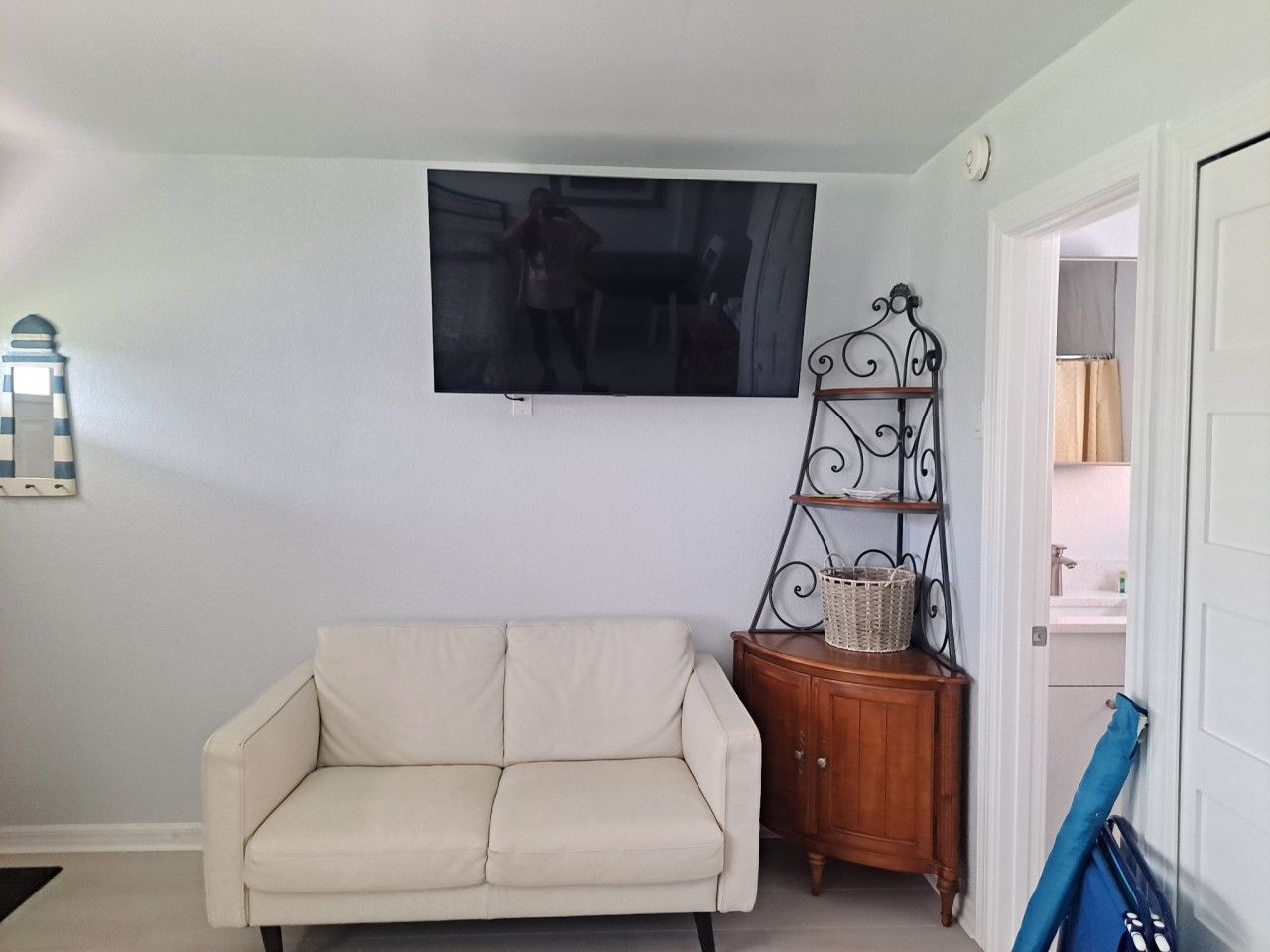 A living room with a sofa, a TV mounted on the wall, a decorative metal stand with a basket, and a door leading to another room.