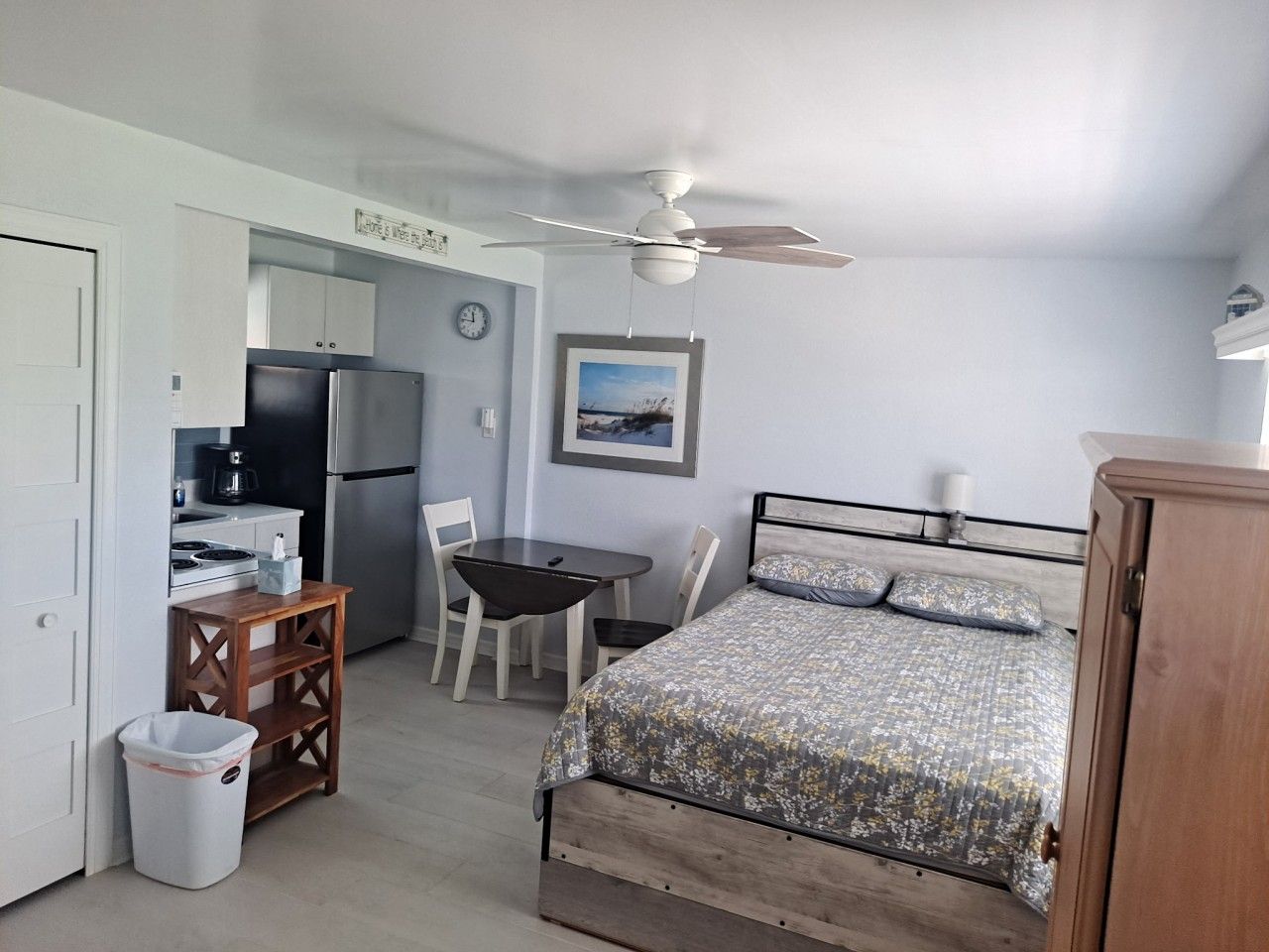A compact room with a bed, a desk, a chair, a trash bin, a ceiling fan, and a refrigerator.