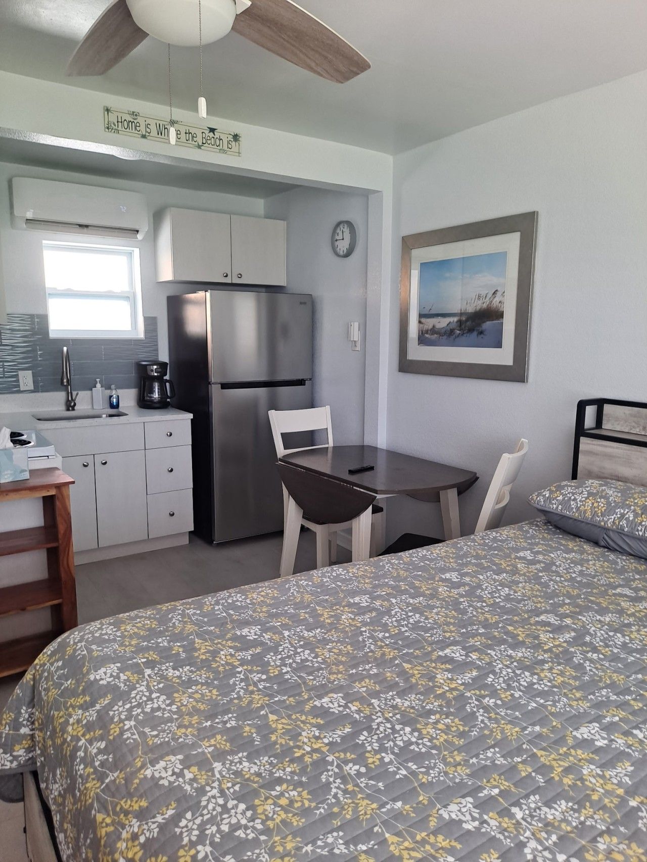 A compact room with a bed, a small table, a chair, a kitchenette, and a framed picture on the wall.