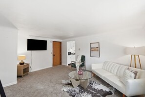 Uma sala de estar moderna com um sofá branco, uma mesa de centro de vidro, uma televisão de tela plana, um armário de madeira e uma porta que dá para uma cozinha.