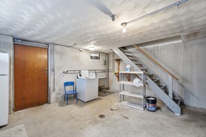 Um porão com porta de madeira, uma cadeira azul, uma máquina de lavar branca, escada de metal e um balde de tinta.