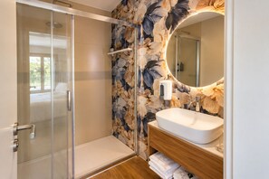A modern bathroom with a glass shower enclosure, a wooden vanity with a sink, and a floral wallpaper design. - House Park (Gandra)