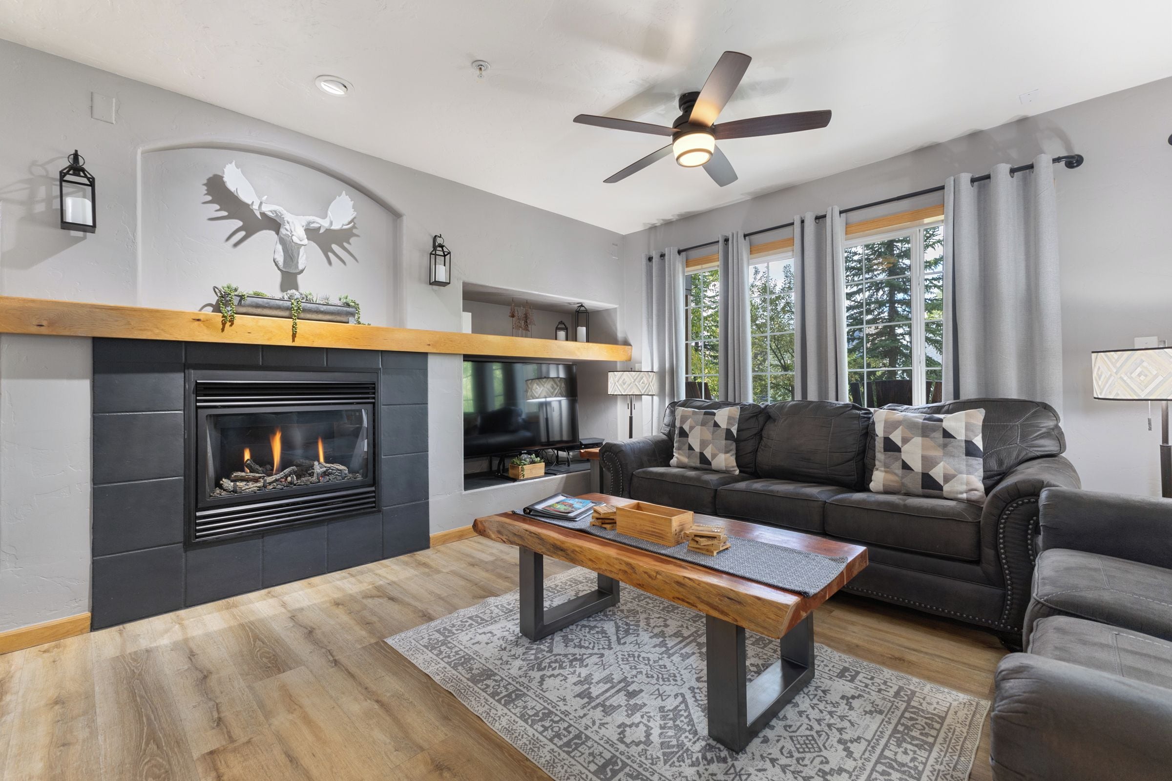 A modern living room with a fireplace, a ceiling fan, and a large window with curtains.
