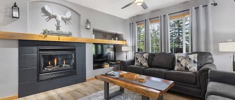 Ein modernes Wohnzimmer mit Kamin, Deckenventilator und großem Fenster mit Vorhängen.