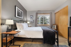 A hotel room with a bed, bedside tables, a desk, a chair, a window with a view of trees, and a wardrobe. - Ground Level | On Bus Route | 3 SmartTVs | Updated (Steamboat Springs)