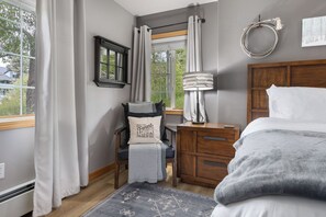 A bedroom with a bed, a nightstand, a lamp, a chair with a cushion, a window with curtains, and a view of greenery outside. - Ground Level | On Bus Route | 3 SmartTVs | Updated (Steamboat Springs)