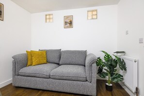 A grey sofa with a mustard-colored pillow, a potted plant, and a radiator in a room with white walls and a wooden floor. - Central London 2-Bedroom Apartment on Leather Lane | Sleeps 6 | (London)