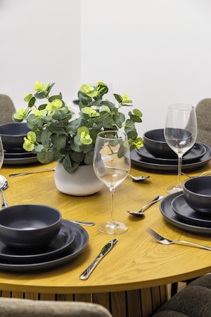 Ein runder Esstisch mit schwarzen Tellern, Besteck und einem klaren Weinglas. In der Mitte steht ein Blumentopf mit grünen Blättern und gelben Blüten.