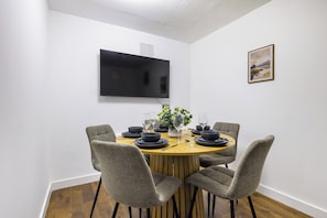 A dining area with a round table, four chairs, a flat-screen TV mounted on the wall, and a framed picture on the wall. - Central London 2-Bedroom Apartment on Leather Lane | Sleeps 6 | (London)