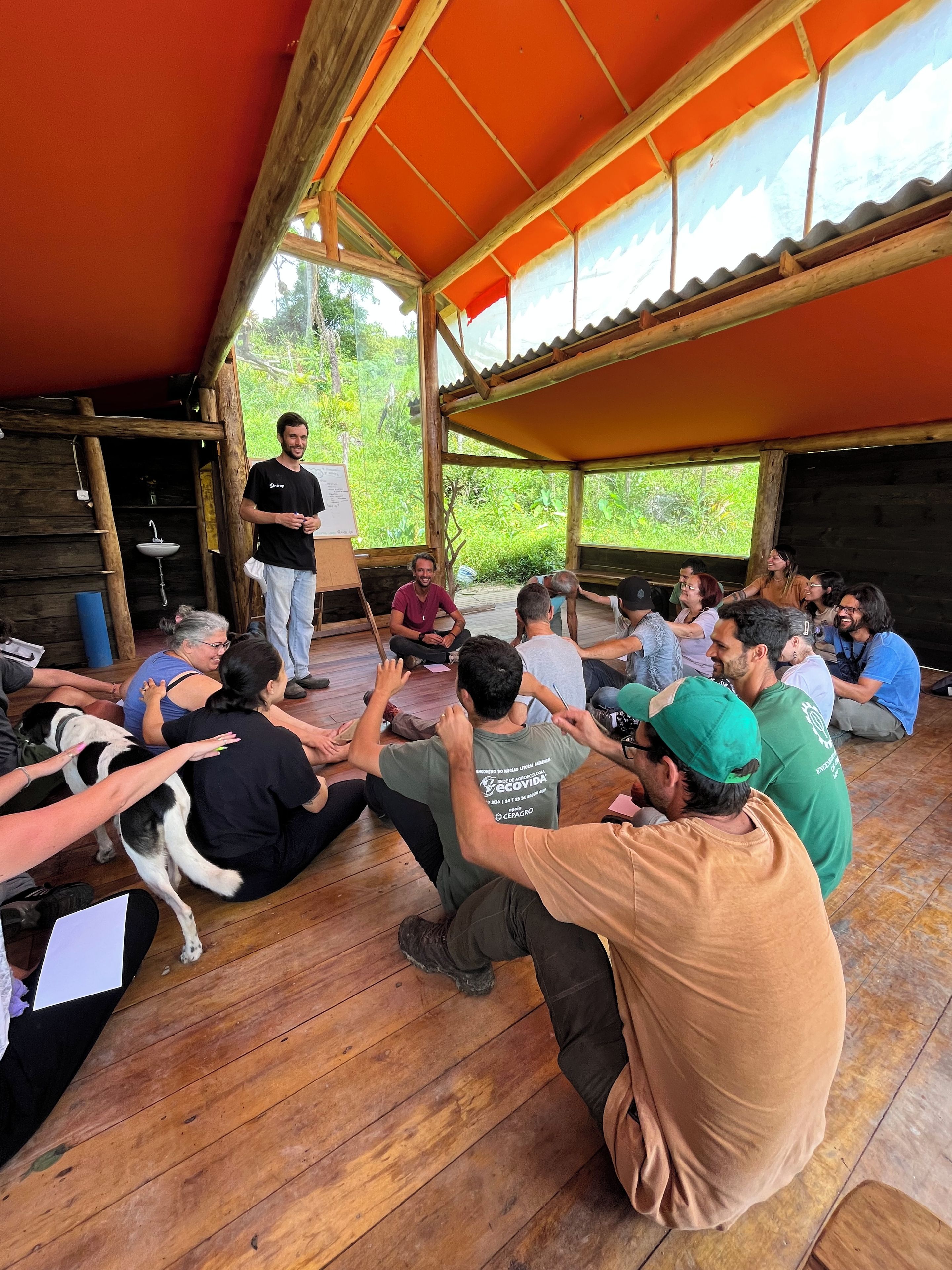 A group of people seated in a wooden-floored room with a red roof, engaged in a meeting or discussion.