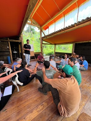 A group of people seated in a wooden-floored room with a red roof, engaged in a meeting or discussion.