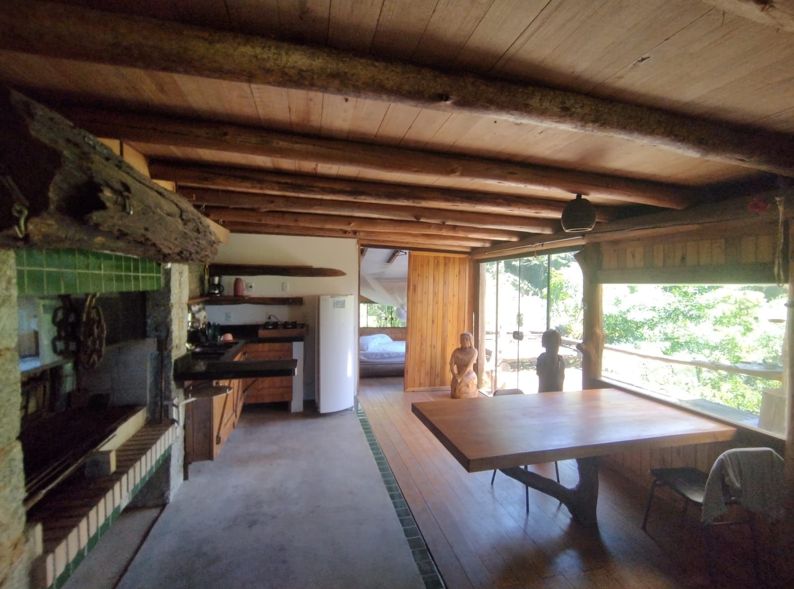 A rustic cabin interior with a wooden ceiling, a kitchen area with a refrigerator, a dining table, and a view through a large window.
