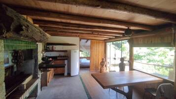 A rustic cabin interior with a wooden ceiling, a kitchen area with a refrigerator, a dining table, and a view through a large window.